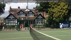 A tennis court with a net, surrounded by a fence, and a building with a sign for Bishops Park Tennis Café in the background. - Stylish self contained patio Garden Apartment in London’s fashionable SW6 (Fulham)