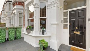 A traditional white townhouse with a black front door, a small porch with potted plants, and a brick facade. - Stylish self contained patio Garden Apartment in London’s fashionable SW6 (Fulham)