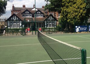 Un court de tennis avec un filet, entouré d’une clôture, et un bâtiment avec une enseigne indiquant le « Bishops Park Tennis Café » en arrière-plan.