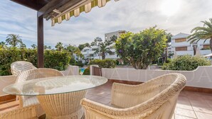 Un espace extérieur aménagé, abrité et doté de chaises en osier et d’une table en verre, offrant une vue sur une piscine et des bâtiments.