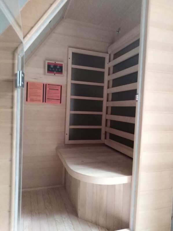 A small, enclosed sauna room with a bench and a door with horizontal slats. There are two informational signs on the wall.