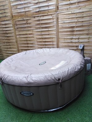 A round, beige inflatable hot tub with a control panel on the right side, placed on artificial grass against a wooden wall.