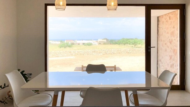 A dining area with a glass table and chairs, two hanging lights, and a view of a landscape through a large window. - Renovated house in Porto Saler (FORMENTERA)