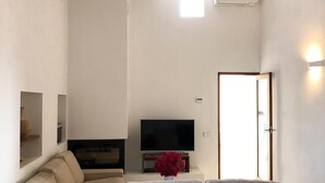 A modern living room with a grey sectional sofa, a flat-screen TV on a stand, and a small red flower vase. - Renovated house in Porto Saler (FORMENTERA)