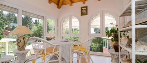 A room with a wooden ceiling, white wicker furniture, and large windows offering a view of greenery.