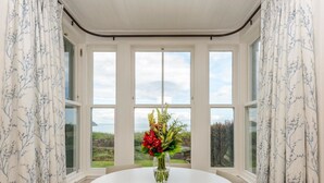Ein runder Tisch mit einem Blumenstrauß in einem Eckzimmer mit großen Fenstern und weißen Vorhängen.