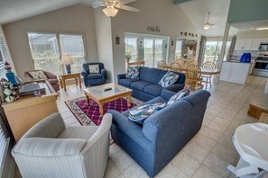 A spacious living room with a blue sofa, a white armchair, and a wooden coffee table. There is a dining area with a table and chairs, and a kitchen with white cabinets and appliances.