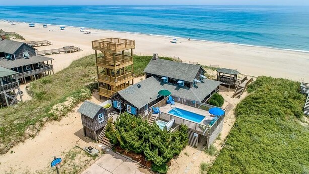 A beachfront property with multiple wooden structures, including a multi-level tower, a pool, and several beachside buildings. - The Beach House | Oceanfront | Dog Friendly, Private Pool, Hot Tub | Nags Head (Nags Head)