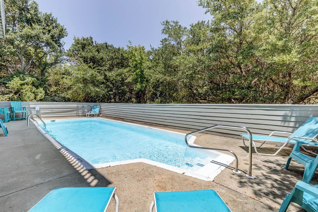 A rectangular outdoor swimming pool with a metal ladder and a white edge. There are blue lounge chairs and a concrete deck surrounding the pool.