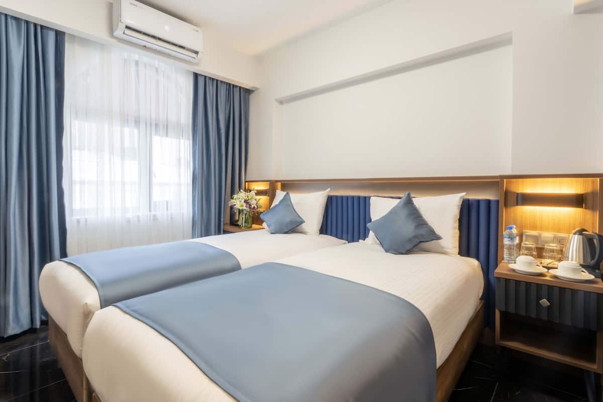 A hotel room with two beds, a nightstand with a lamp, and a window with curtains.