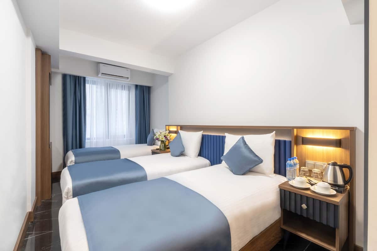 A hotel room with two beds, a nightstand with a kettle, and a window with curtains.