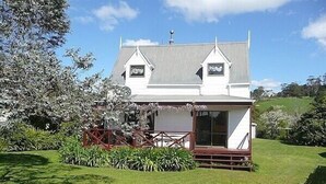 A two-story house with a white exterior, a grey roof, and a small porch with a wooden railing. - Cute Cottage - Cooks Beach Bach (Cooks Beach)