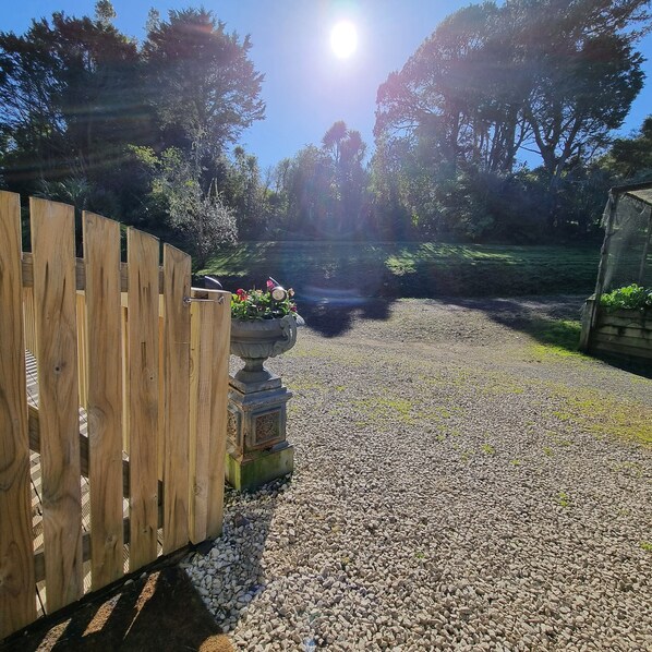 Un chemin de gravier mène à une clôture en bois et à un jardin agrémenté d’un vase décoratif et de fleurs.
