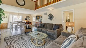 A spacious living room with a large sofa, a coffee table, and a dining area with a table and chairs. A staircase with wooden railings is visible in the background. - Splash and Go | 1135 ft from the beach | Private Pool, Hot Tub | Nags Head (Nags Head)