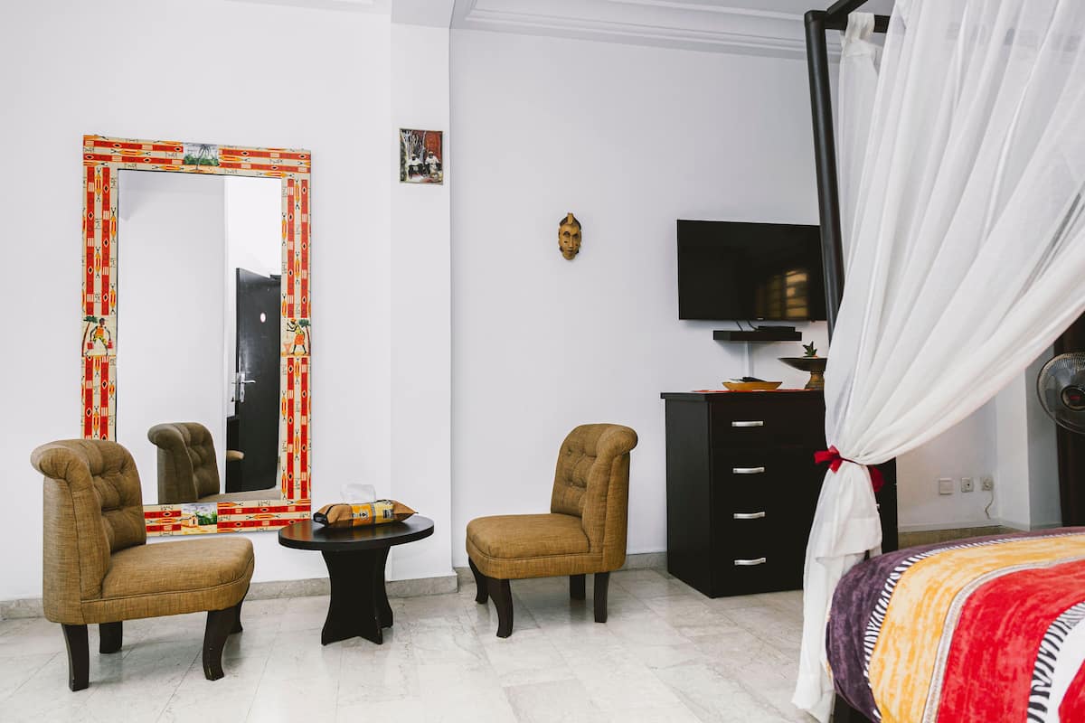Una habitación con una cama, dos sillones, una mesita, un televisor, un armario y un espejo con un marco decorativo.