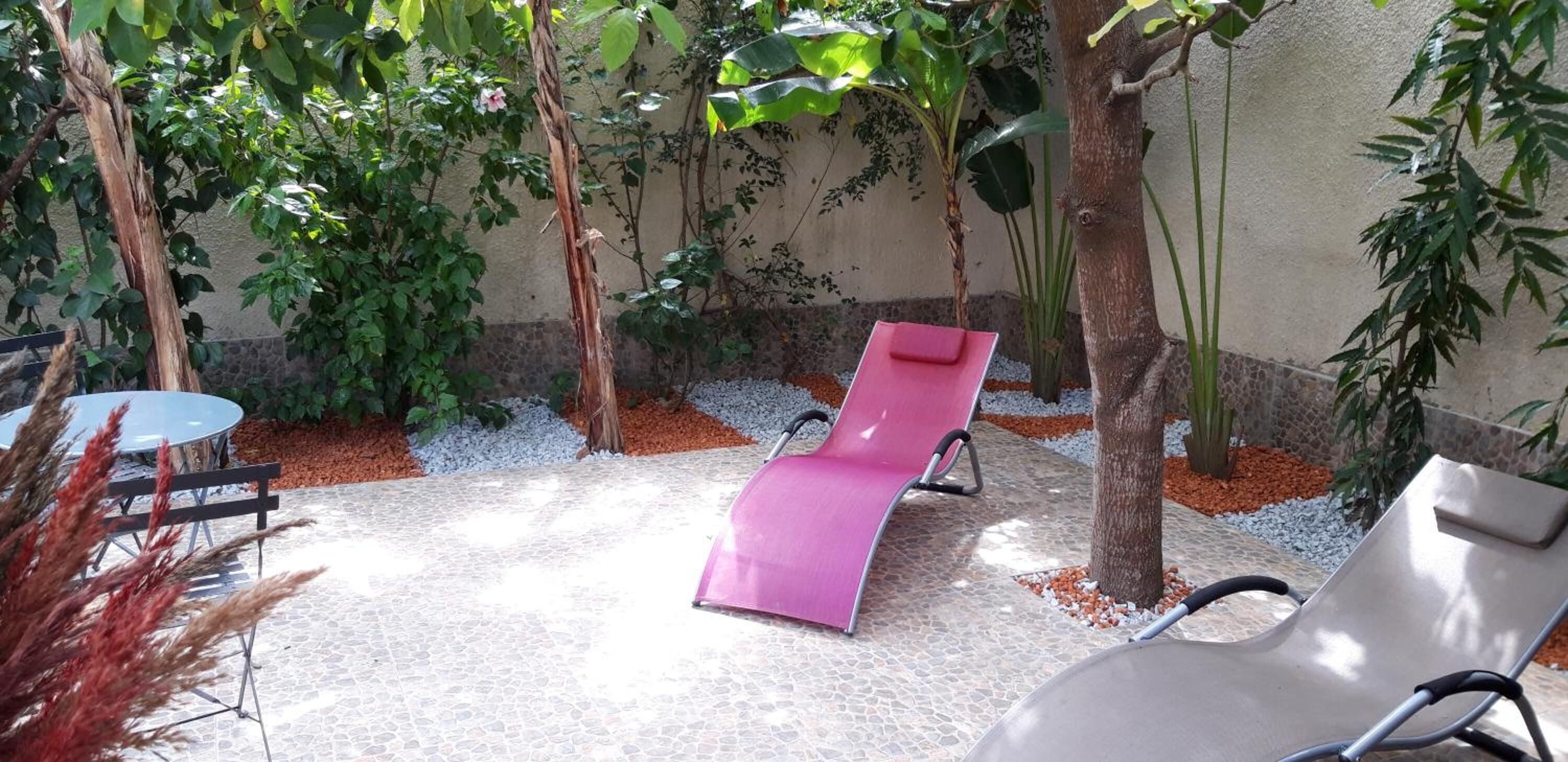 Un patio con mesa, dos sillones de relax y un pequeño jardín con plantas y un árbol.