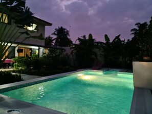 Una piscina con superficie de agua cristalina, rodeada de plantas tropicales y un edificio con toldo rosado.