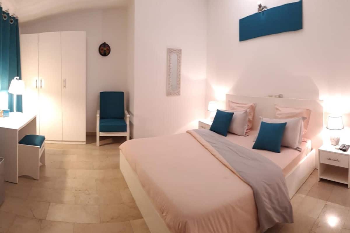 Habitación de hotel con cama, mesita de noche, lámpara, silla, escritorio, armario y televisor.