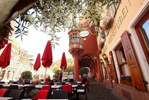Un café al aire libre con sombrillas rojas y mesas, una torre del reloj y un letrero que indica Hotel Oberkirchner Restaurant.