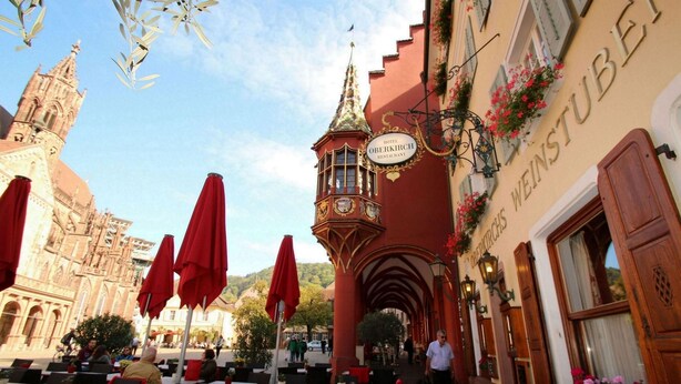 Een terras met tafels en rode parasols, een gebouw met een bord waarop staat 'Hotel Oberkirch Restaurant', en een kerk op de achtergrond.