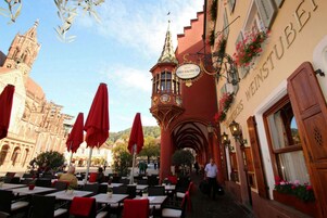 Un área de café al aire libre con mesas y sombrillas rojas, un edificio con un letrero que dice "Hotel Oberkirch Restaurant", y una iglesia al fondo.