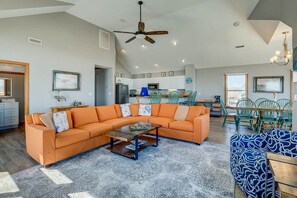A spacious living area with a large orange sectional sofa, a coffee table, and a blue patterned sofa. There is a dining area with green chairs and a kitchen in the background. - Krosnest | Oceanfront | Private Pool, Hot Tub | Nags Head (Nags Head)