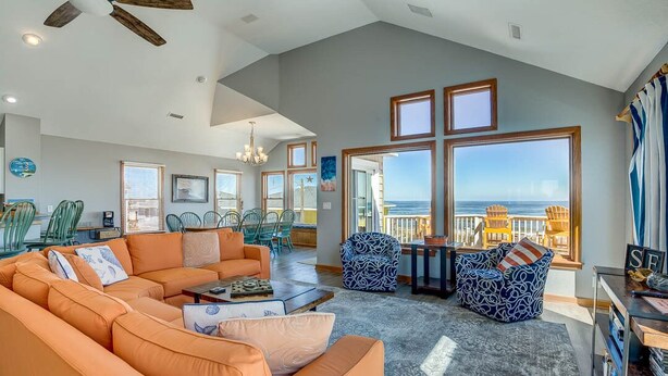 A spacious living room with a large orange sectional sofa, a coffee table, and patterned chairs. There are large windows offering a view of the ocean. - Krosnest | Oceanfront | Private Pool, Hot Tub | Nags Head (Nags Head)