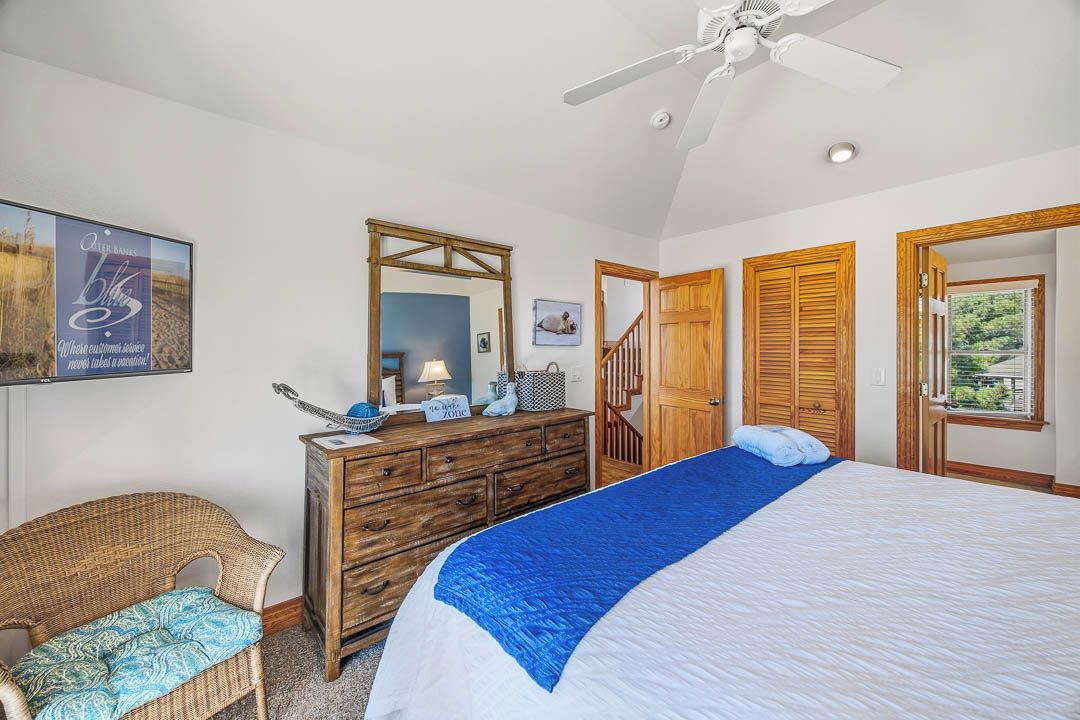 A bedroom with a bed, a dresser, a chair, a ceiling fan, and a door leading to another room.