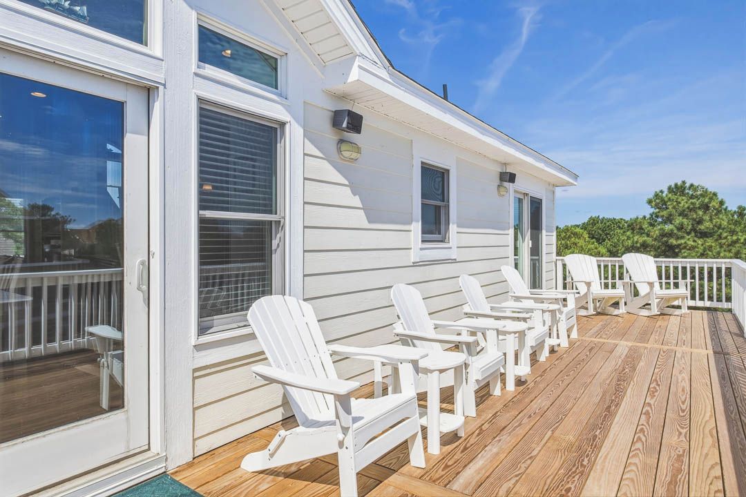 A deck with white Adirondack chairs, a wooden deck, and a white building with windows and a door.