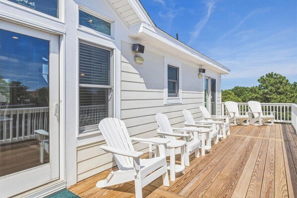 Una terraza con sillas Adirondack blancas, una cubierta de madera y un edificio blanco con ventanas y una puerta.