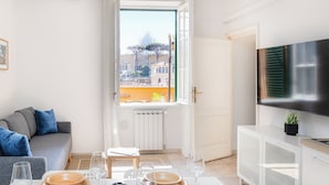 A modern living room with a dining table set for four, a sofa, a TV, and a window with a city view. - Rental In Rome Sistina View Terrace Apartment (Rome)