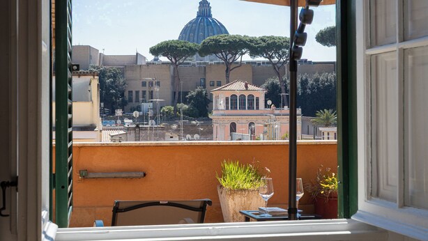 A balcony with a view of a dome and trees, a table with a glass, and a potted plant. - Rental In Rome Sistina View Terrace Apartment (Rome)