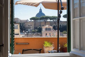 A balcony with a view of a dome and trees, a table with a glass, and a potted plant. - Rental In Rome Sistina View Terrace Apartment (Rome)