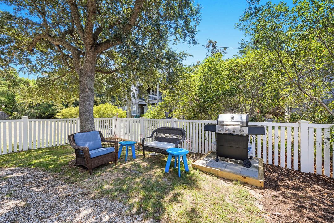 A backyard with a grill, two blue stools, a wicker chair, and a white picket fence.