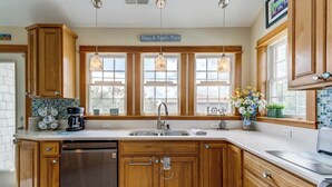 A kitchen with wooden cabinets, a stainless steel dishwasher, a double sink, pendant lights, and a window with a view. - Loafing Around In Duck | 1250 ft from the beach | Private Pool, Hot Tub | Duck (Duck)