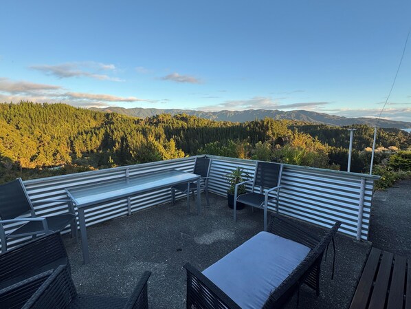 Une terrasse dotée de rampes en métal, d’un banc et de deux chaises, offrant une vue sur une chaîne de montagnes boisée.