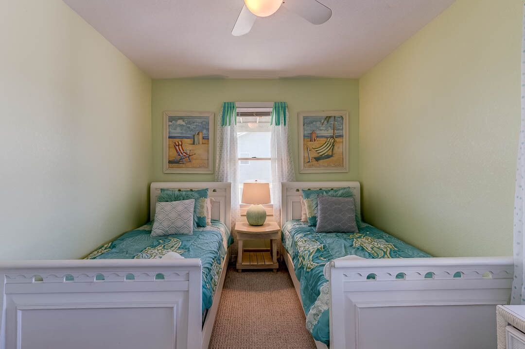 A room with two beds, a ceiling fan, a window with curtains, and two wall-mounted paintings.