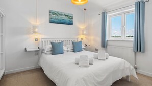 A neatly made bed with white linens and blue pillows, a painting of a coastal scene, and a window with blue curtains.