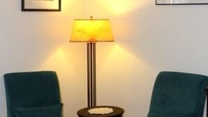 Ein Zimmer mit zwei türkisblauen Sesseln, einem kleinen runden Tisch mit einer Lampe und gerahmten Bildern an den Wänden.