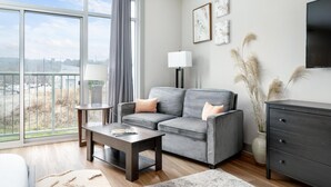 A modern living room with a grey sofa, a coffee table, a TV, and a view of the city through large windows. - Studio King Suite w/ Kitchen | 200 (Asheville)