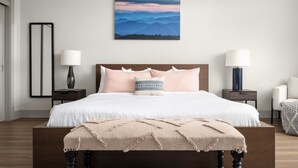 A bedroom with a large bed, two bedside tables, a nightstand, a chair, and a painting on the wall. - Studio King Suite w/ Kitchen | 200 (Asheville)