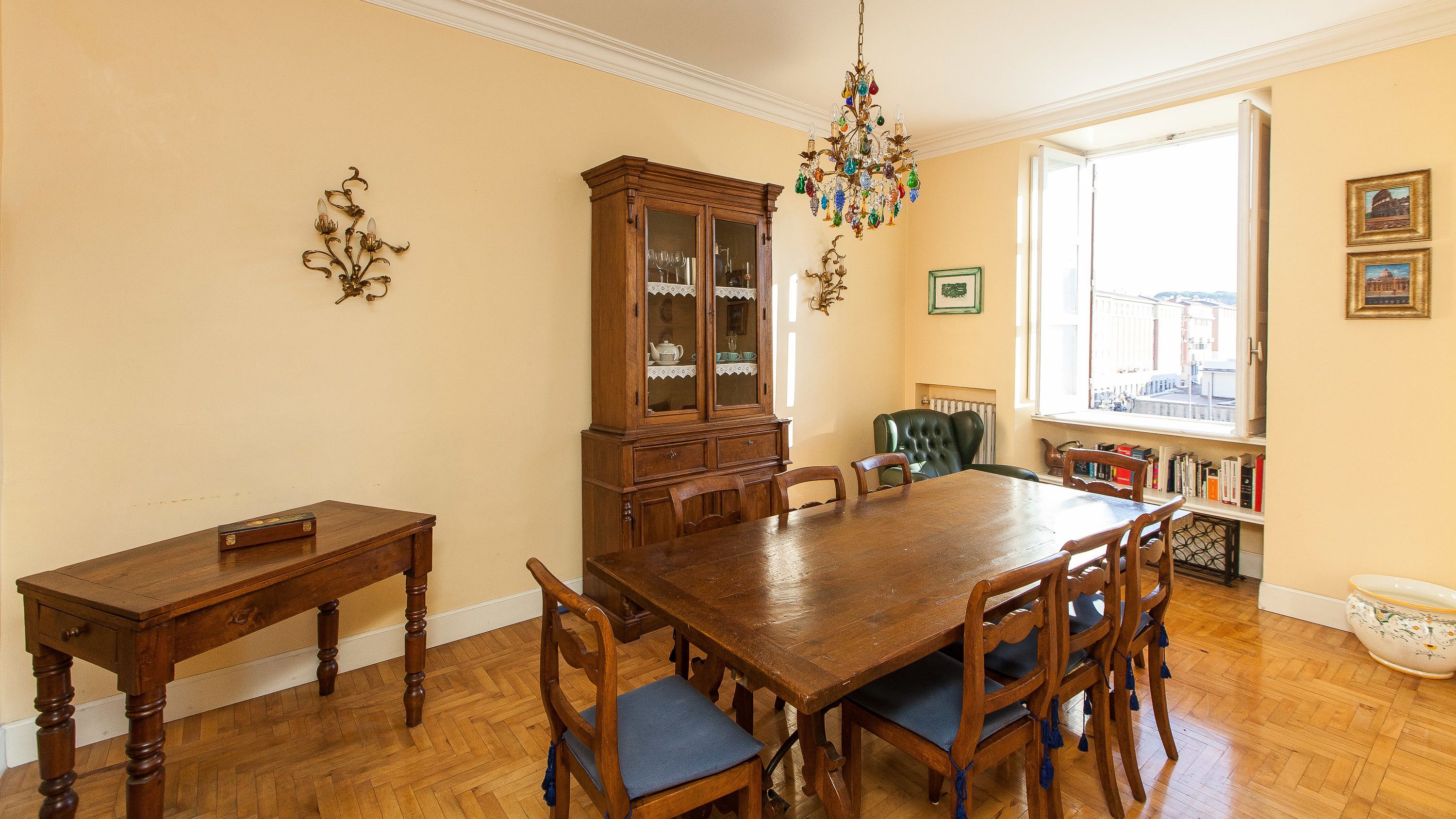 A dining room with a wooden table and chairs, a cabinet with glass doors, a chandelier, and a window with a view of buildings.