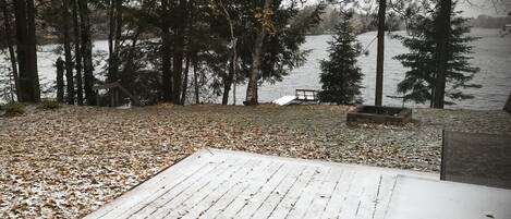 A wooden deck with a bench and a table, overlooking a lake with trees and a dock.