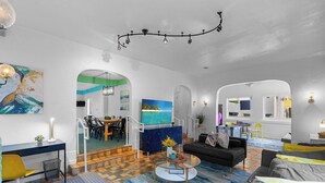 Ein modernes Wohnzimmer mit Sofa, Couchtisch und Fernseher. Es gibt einen Essbereich mit Stühlen und Tisch. Der Raum ist blau und weiß gestaltet.