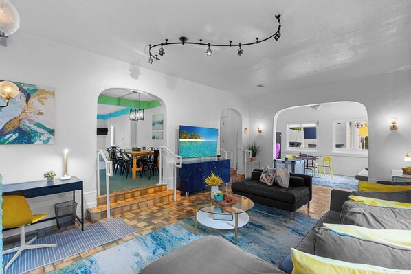 A modern living room with a sofa, coffee table, and a TV. There is a dining area with chairs and a table. The room has a blue and white color scheme.