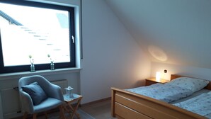 Ein Schlafzimmer mit einem Bett, einem Sessel, einem kleinen Tisch und einem Fenster mit Blick auf Gebäude.