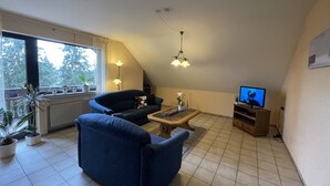 Ein Wohnzimmer mit einer Couch, einem Couchtisch, einem Fernseher und einem Fenster mit Pflanzen.
