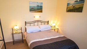 Un lit bien fait, recouvert d’une couverture bleu foncé, avec deux lampes de chevet et deux tableaux représentant une scène de plage, accrochés au mur.