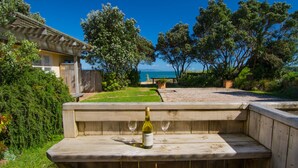 Un espace extérieur aménagé avec des sièges en bois, une bouteille de vin et un verre, entouré de verdure et offrant une vue sur la mer.