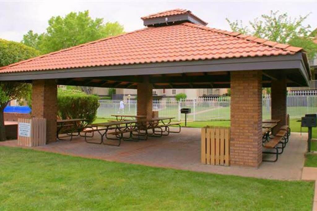 A covered picnic area with picnic tables and benches, surrounded by a brick structure and a fenced sports field in the background.